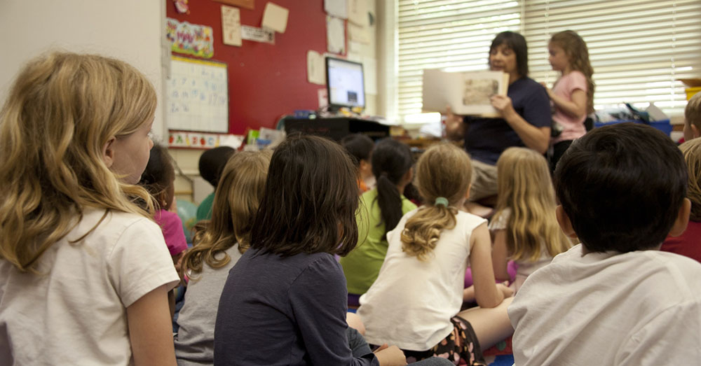 children seated on the floor in a classroom, listening to a teacher