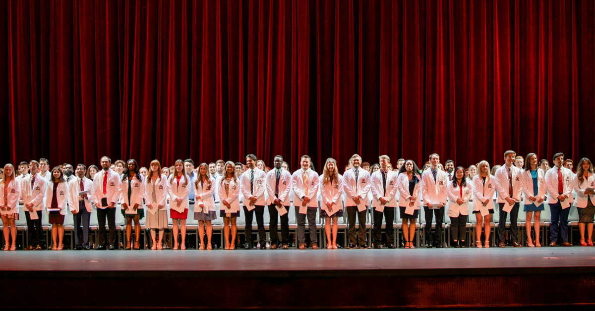 Large group of TTUHSC medical students stand on stage wearing their white coats.