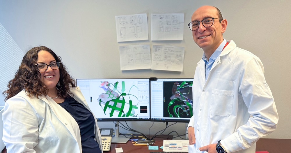 two scientists in lap coats stand in front of a computer screen
