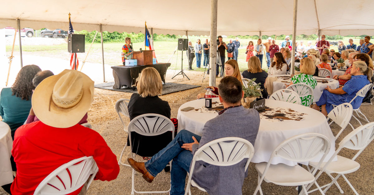 Large group of people sits outside at tables under a tent while a woman speaks at a podium.