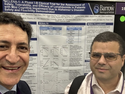 two men smile in a selfie photo in front of a research poster at a conference