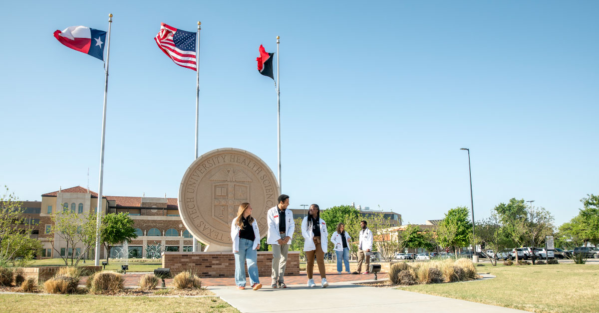 Five medical students wear white coats and stand outside in front of the TTUHSC seal.