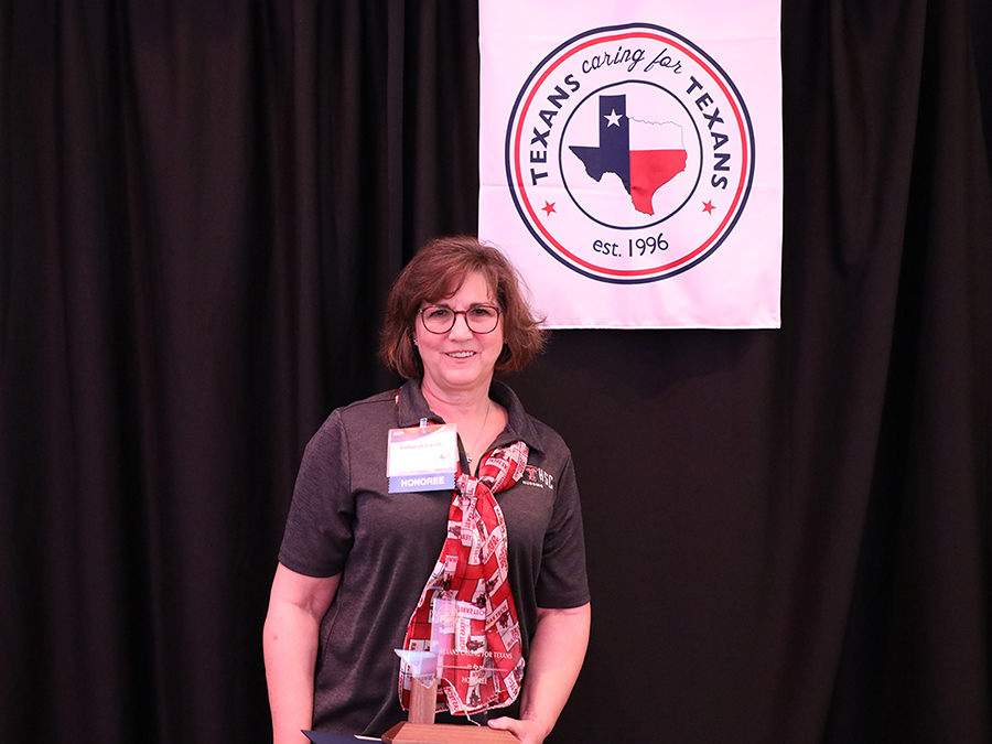 Female TTUHSC nursing professor holds a trophy and stands under a banner that reads, "Texans Caring for Texans."