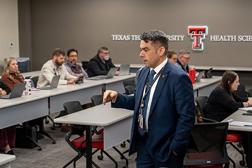Man wears suit and leads a meeting full of people.