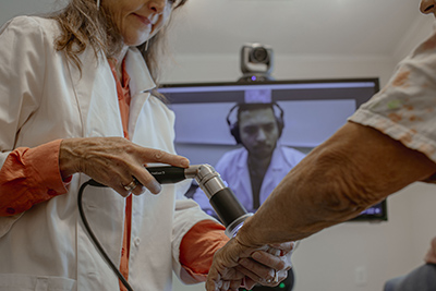 Female clinician holds a male patient's arm while a male physician watches via telehealth on a computer screen.