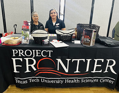 Two woman sit behind a table with a table cloth decorated with the words, "ProjectFRONTIER."