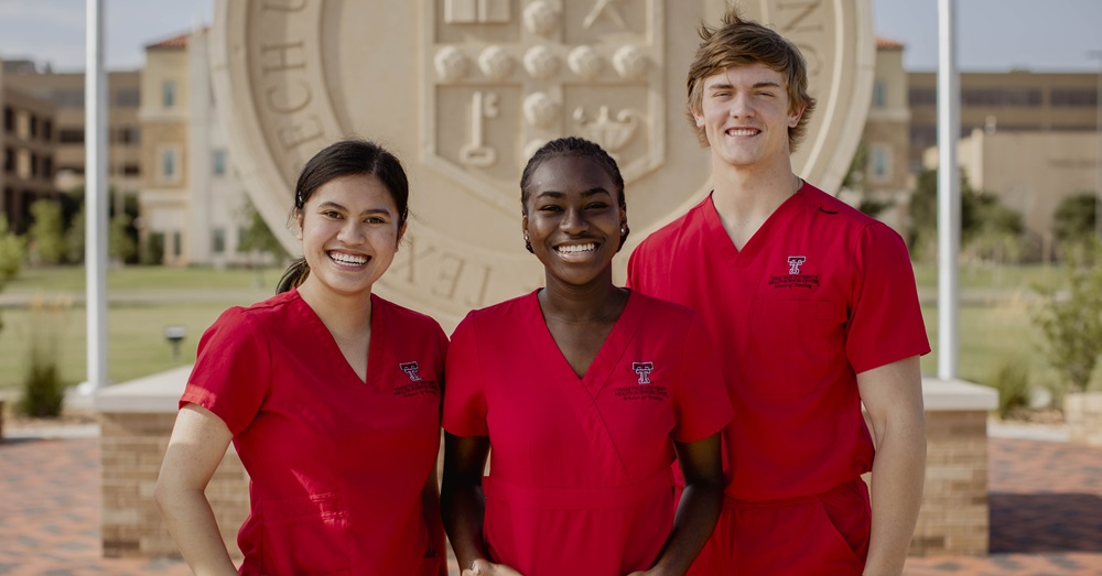 three students in red scrubs smile in front of the TTUHSC seal in Lubbock