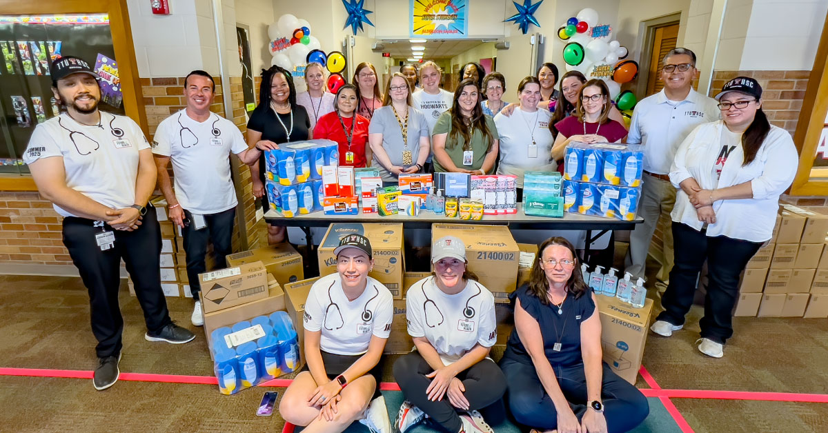 Large group of TTUHSC faculty and students stand around a table full of school supply donations.