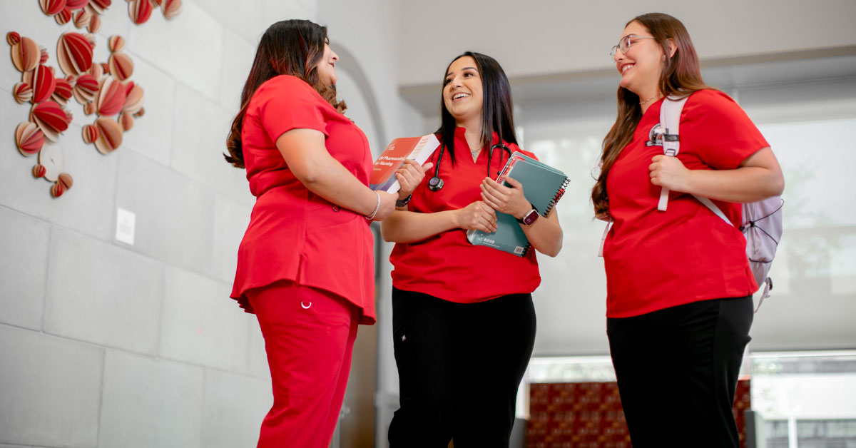 Three nursing students wearing red scrubs stand and talk.