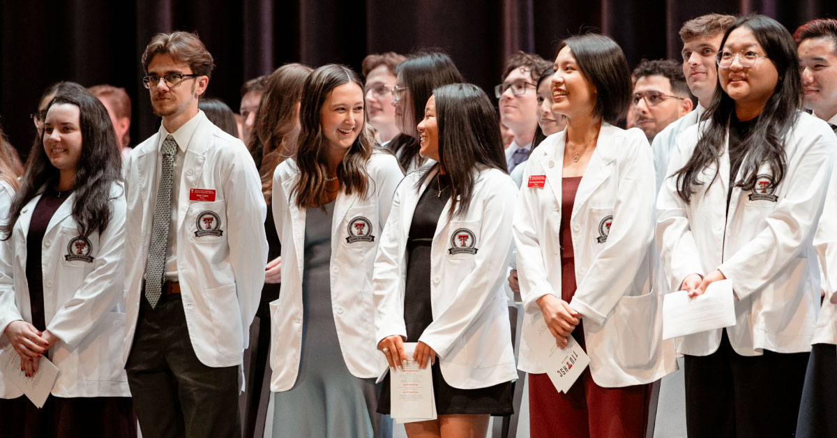 Large group of medical students stands on stage while wearing their white coats.
