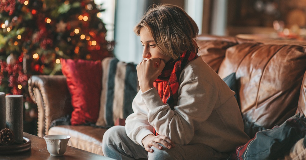 woman sitting on a couch with a cup of coffee looking sad with a christmas tree behind her