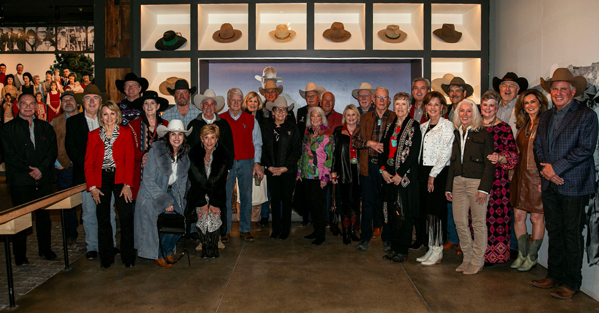 Large group of donors pose for a group photo in front of a wall full of cowboy hats.