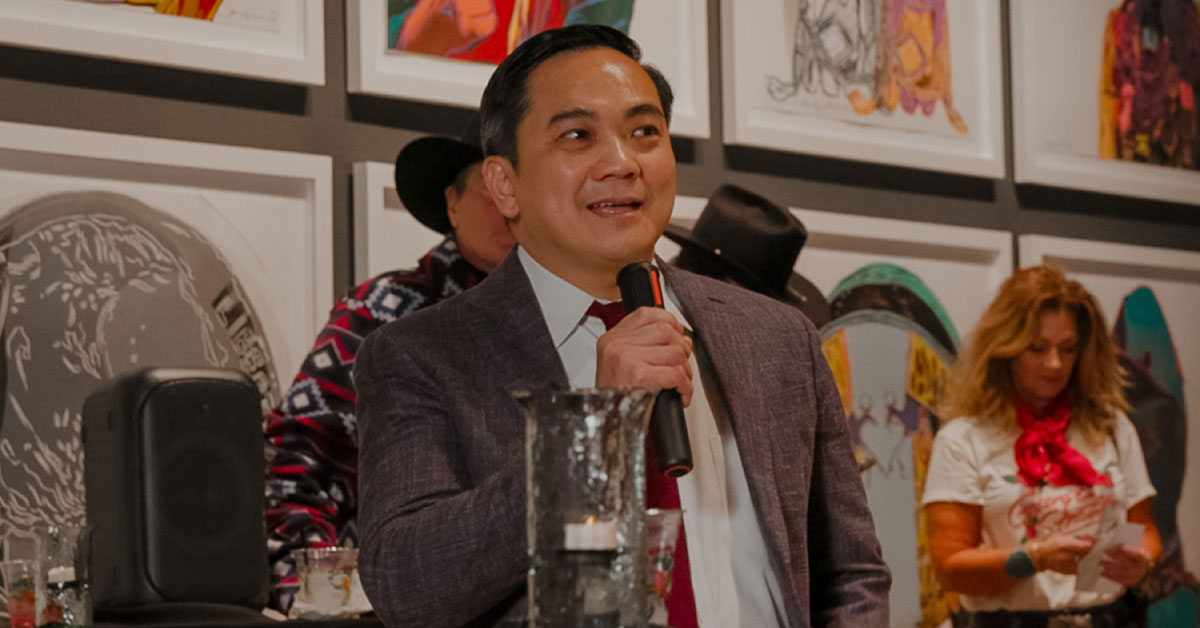 Man in suit stands and speaks to a room full of people.