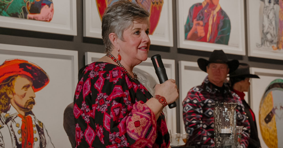 Woman in dress stands and speaks to a room full of people.