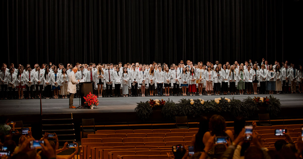 Large group of medical students wearing white coats on stage.