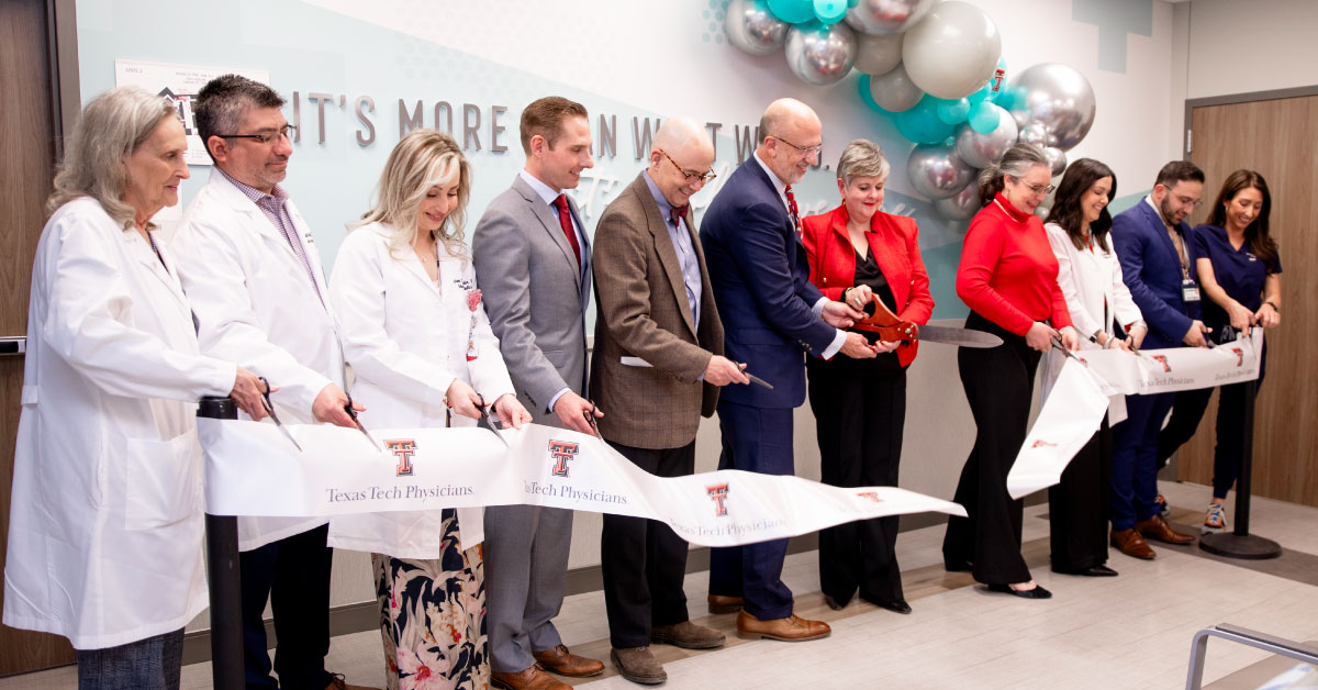 Long line of doctors and administrators cut a ribbon with a large pair of scissors.