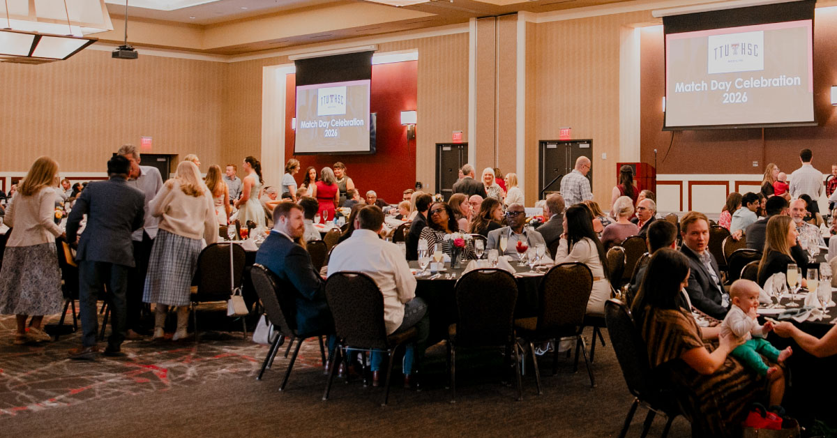 Many people fill several banquet tables in a large room with "Match Day Celebration" projected on two large screens.