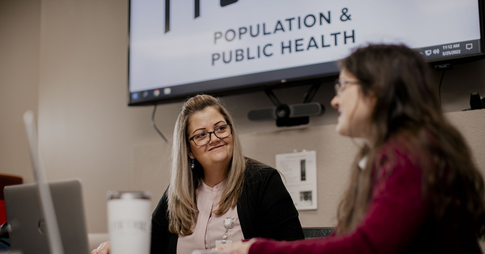 two women talking at a table with a screen behind them that says "population and public health"