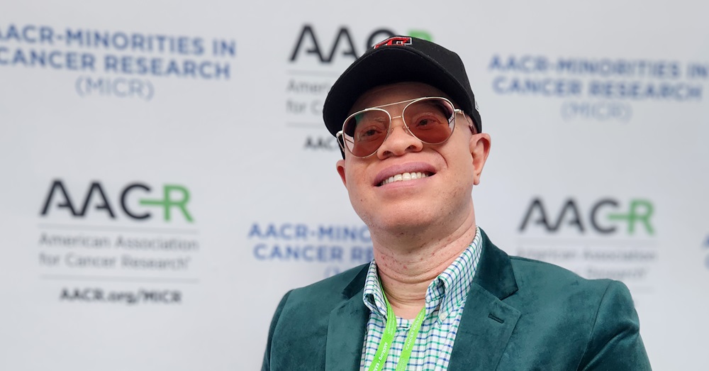 man smiling in front of a backdrop wearing glasses and a hat