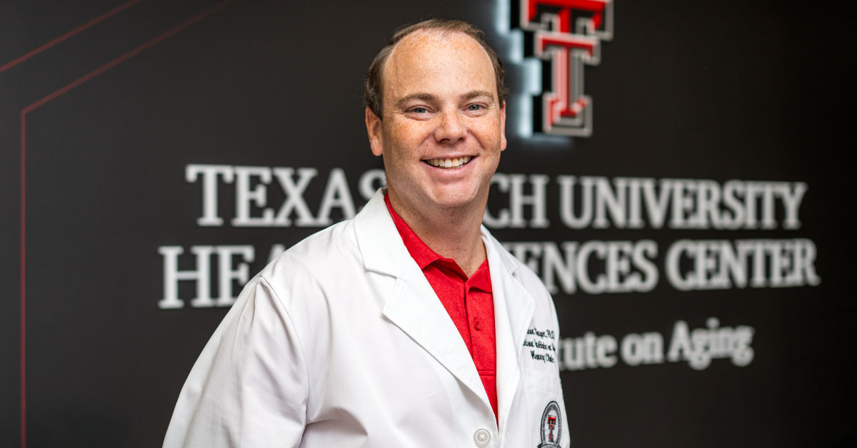 Jonathan Singer, Ph.D., wears a white coat and stands in front of a wall decorated with the TTUHSC Garrison Institute on Aging logo.