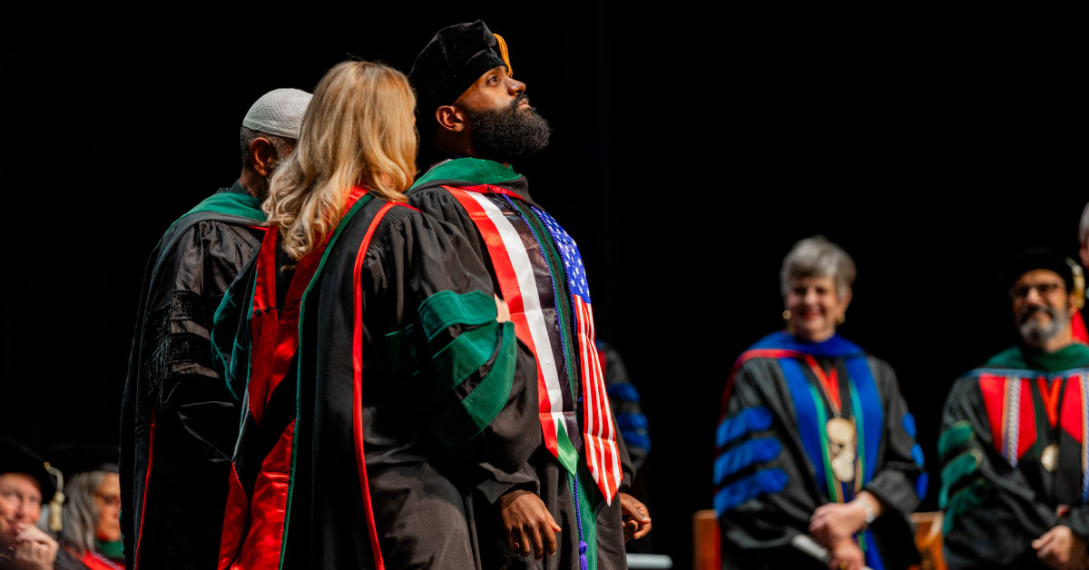 Man wears graduation regalia as he is hooded on stage and other people watch.