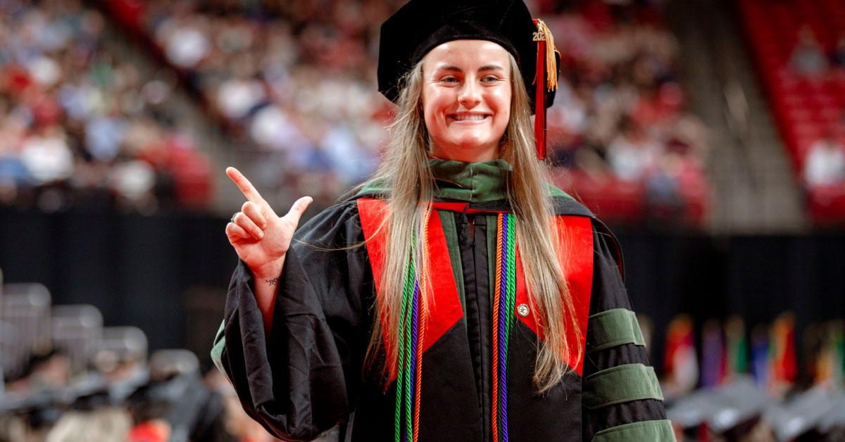 Female TTUHSC student wears her graduation cap and gown and holds up the "guns up" hand sign.